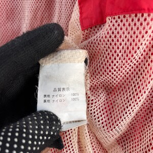 Puede incluir: Una etiqueta blanca con texto negro que dice "商品表示"  "表地 ナイロン 100%" "裏地 ナイロン 100%"  La etiqueta est&aacute; unida a una tela de malla roja y blanca.