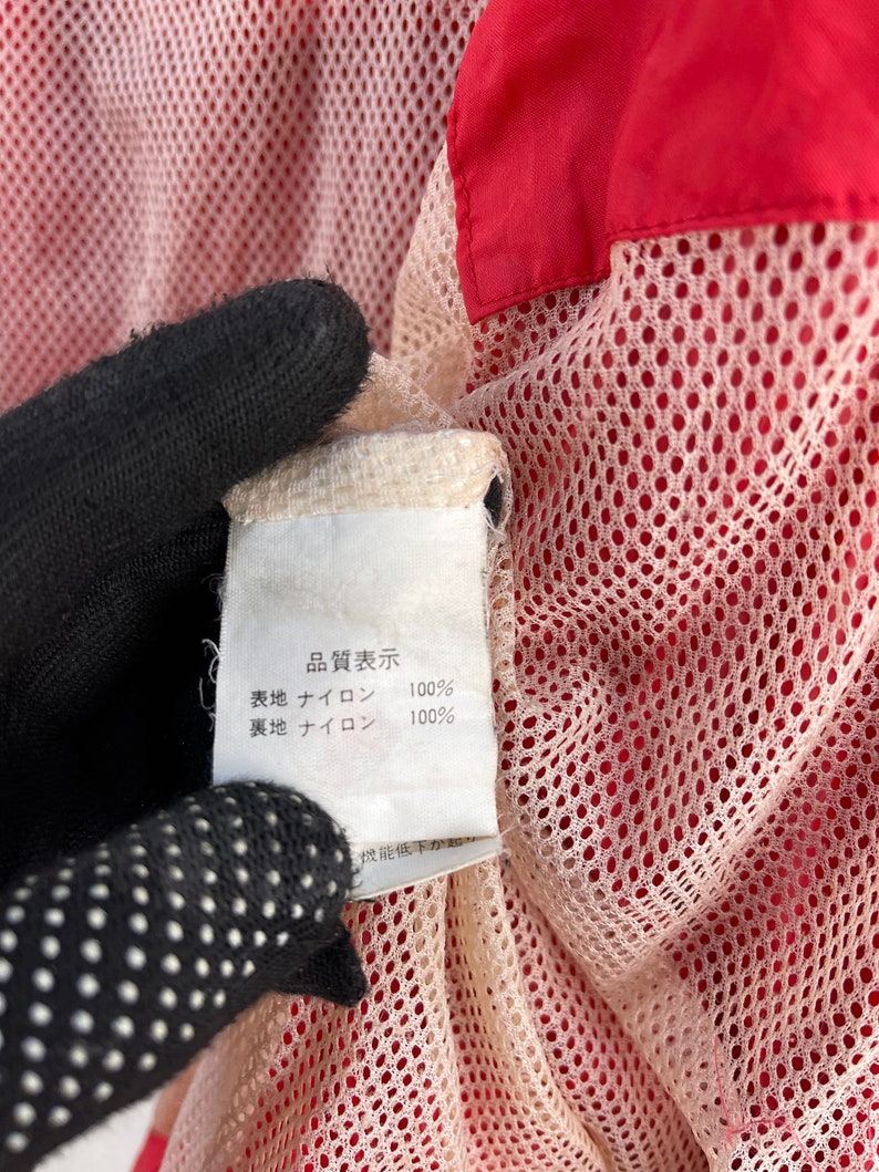 Puede incluir: Una etiqueta blanca con texto negro que dice "商品表示"  "表地 ナイロン 100%" "裏地 ナイロン 100%"  La etiqueta est&aacute; unida a una tela de malla roja y blanca.