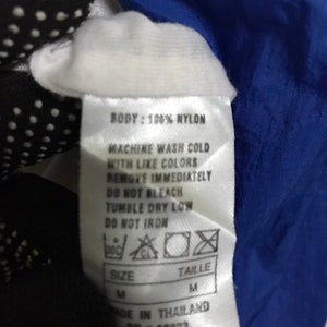 Op de afbeelding: Een wit label met zwarte tekst met details over de wasinstructies voor een kledingstuk van nylon. Het label zegt "BODY: 100% NYLON", "MACHINE WASH COLD WITH LIKE COLORS", "REMOVE IMMEDIATELY", "DO NOT BLEACH", "TUMBLE DRY LOW", "DO NOT IRON", "SIZE M", "TAILLE M", "MADE IN THAILAND", "RN # 56323".