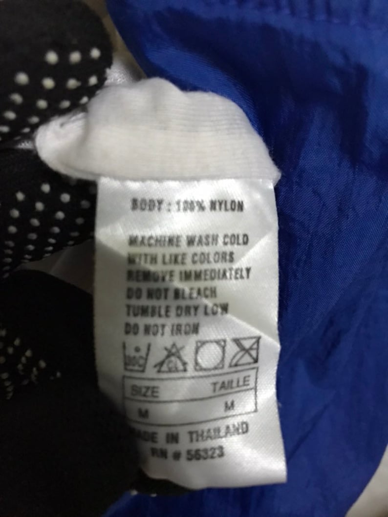 Op de afbeelding: Een wit label met zwarte tekst met details over de wasinstructies voor een kledingstuk van nylon. Het label zegt "BODY: 100% NYLON", "MACHINE WASH COLD WITH LIKE COLORS", "REMOVE IMMEDIATELY", "DO NOT BLEACH", "TUMBLE DRY LOW", "DO NOT IRON", "SIZE M", "TAILLE M", "MADE IN THAILAND", "RN # 56323".