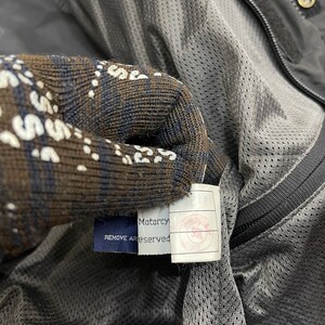 Puede incluir: Una chaqueta negra con forro de malla y una etiqueta azul que dice "Motarcy REMOVE ALL TAGS RESERVED".