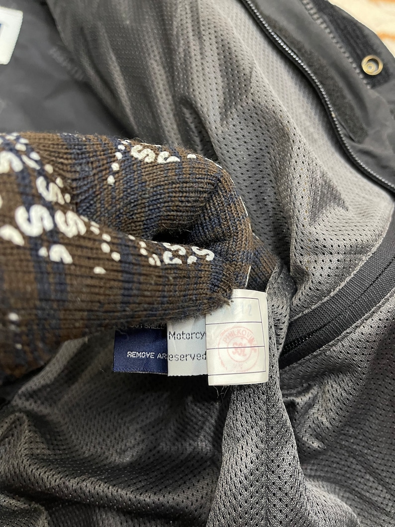 Puede incluir: Una chaqueta negra con forro de malla y una etiqueta azul que dice "Motarcy REMOVE ALL TAGS RESERVED".