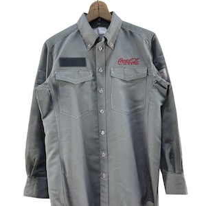 Peut inclure: Une chemise à manches longues à boutons, à rayures, avec un logo Coca-Cola brodé sur la poitrine. La chemise a deux poches poitrine et un patch sur la manche gauche.