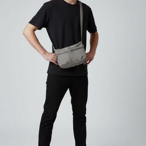 May include: A gray crossbody bag with multiple zippered compartments. The bag is worn over the shoulder. The person in the image is wearing a black t-shirt, black pants, and black sneakers.