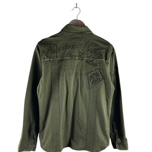 May include: Olive green long-sleeved shirt with buttoned cuffs and a collar, hanging on a wooden hanger. The back of the shirt features embroidered text that reads "Superior Mother Core In The Law" and "Wild Sup Style".