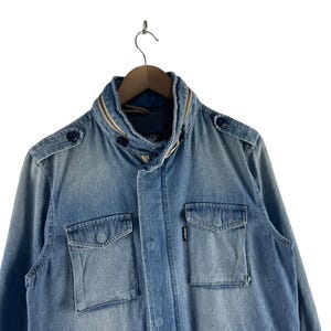 Puede incluir: Chaqueta vaquera azul claro con cuello alzado y dos bolsillos delanteros con solapa. La chaqueta tiene cierre de botones y botones decorativos en los hombros. La chaqueta est&aacute; colgada de una percha de madera.