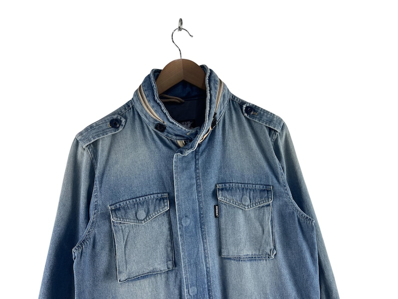 Puede incluir: Chaqueta vaquera azul claro con cuello alzado y dos bolsillos delanteros con solapa. La chaqueta tiene cierre de botones y botones decorativos en los hombros. La chaqueta est&aacute; colgada de una percha de madera.