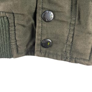 May include: Close-up of an olive green jacket with two black buttons. The buttons are embossed with the word "AVIREX" and the letters "U.S.A." The jacket has a ribbed knit cuff.