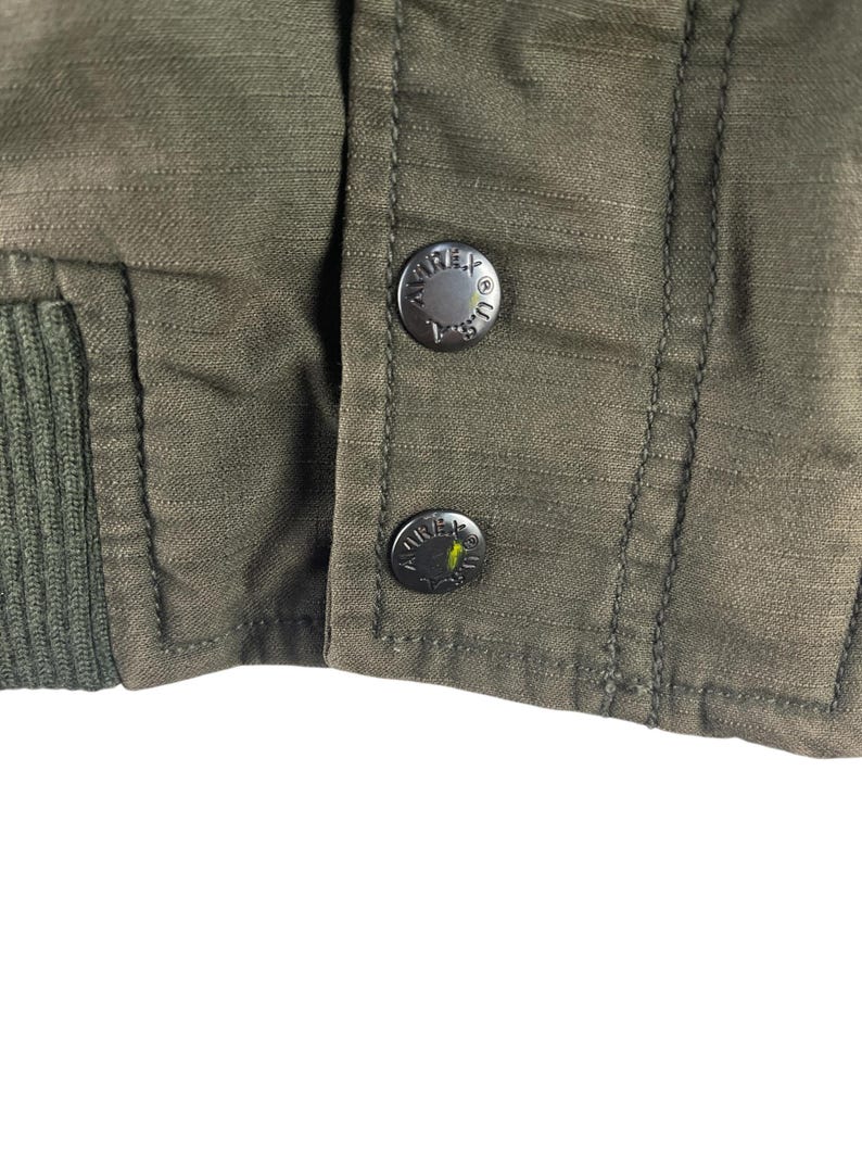 May include: Close-up of an olive green jacket with two black buttons. The buttons are embossed with the word "AVIREX" and the letters "U.S.A." The jacket has a ribbed knit cuff.
