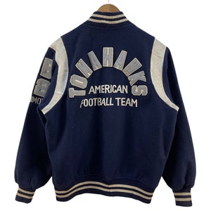 May include: A navy blue varsity jacket with white and tan striped ribbing. The back of the jacket features a large embroidered patch that reads "Tomahawks American Football Team".