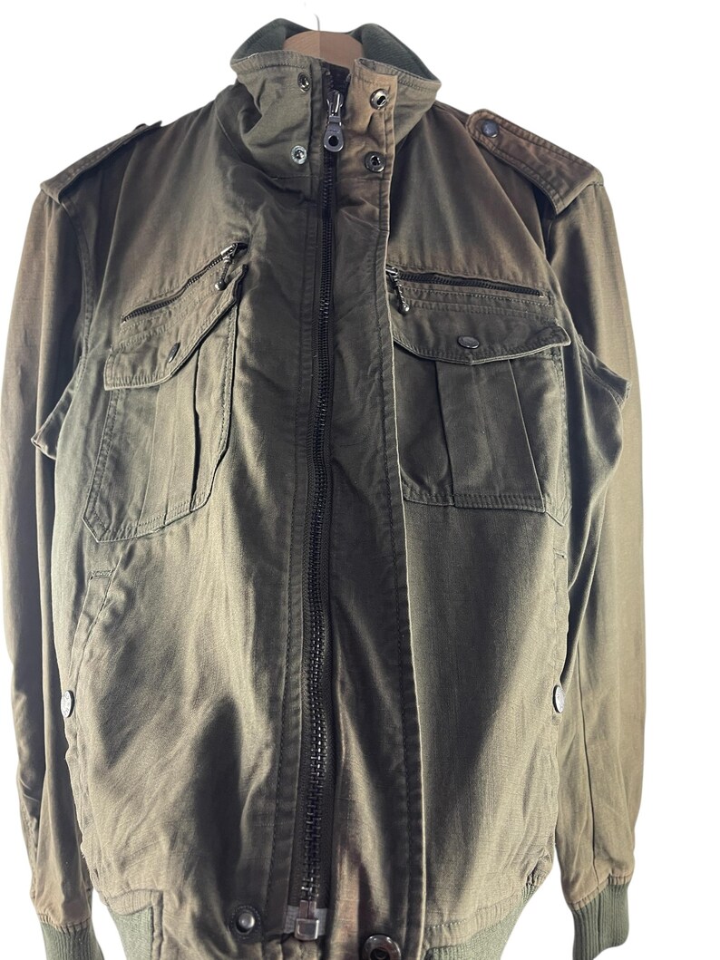 May include: Olive green bomber jacket with a zip-up front, multiple pockets, and a ribbed collar and cuffs. The jacket has a military-inspired design with shoulder epaulets and a relaxed fit. The fabric appears to be a durable, woven material.
