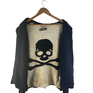 May include: Black open-front sweater with a large skull and crossbones design on the front. The sweater is hanging on a wooden hanger against a white background. The skull and crossbones are black.