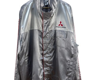 May include: A silver jacket with red accents and a Mitsubishi Motors logo on the front. The jacket has a zipper closure and two pockets.