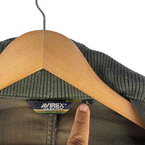 May include: Olive green varsity jacket with a ribbed collar, hanging on a wooden hanger. The Avirex Varsity label is visible, along with a size "L" tag. The jacket is made of a durable fabric.