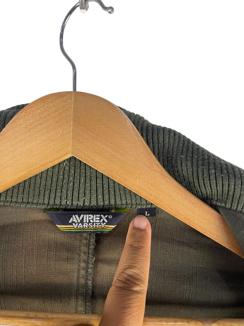 May include: Olive green varsity jacket with a ribbed collar, hanging on a wooden hanger. The Avirex Varsity label is visible, along with a size "L" tag. The jacket is made of a durable fabric.