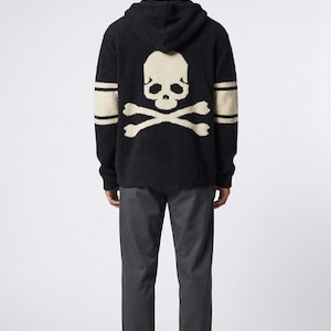 May include: Black hooded sweater with a large skull and crossbones design on the back. The sweater has cream-colored accents on the sleeves and skull. The model is wearing dark gray pants and white sneakers.