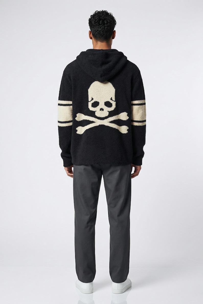 May include: Black hooded sweater with a large skull and crossbones design on the back. The sweater has cream-colored accents on the sleeves and skull. The model is wearing dark gray pants and white sneakers.