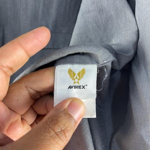 Puede incluir: Primer plano de una etiqueta de prenda gris con la marca AVIREX&reg; y un emblema dorado con alas estilizadas. La etiqueta est&aacute; adherida a una tela gris, con la textura de la tela visible.