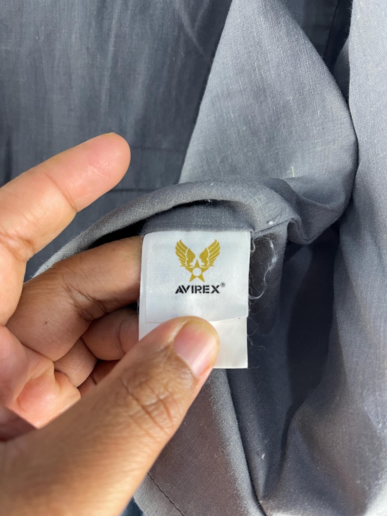Puede incluir: Primer plano de una etiqueta de prenda gris con la marca AVIREX&reg; y un emblema dorado con alas estilizadas. La etiqueta est&aacute; adherida a una tela gris, con la textura de la tela visible.