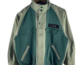 May include: A two-tone jacket in olive green and teal. The jacket features a stand-up collar, button closures, and two front pockets. A small patch with Japanese text is on the upper chest. The jacket is hanging on a wooden hanger.