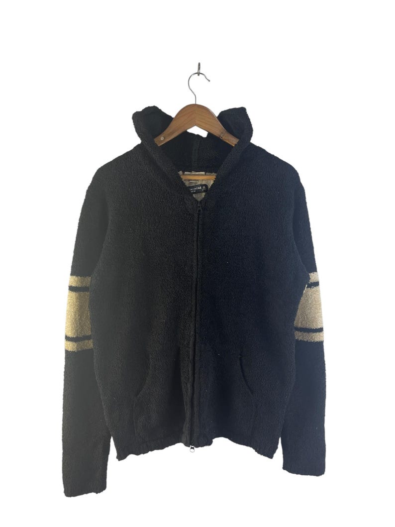 May include: Black zip-up hooded sweater with tan stripes on the sleeves. The sweater has a textured, fuzzy appearance, a hood, and two front pockets. The sweater is hanging on a wooden hanger.