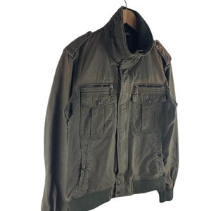 May include: Olive green bomber jacket with a high collar and multiple front pockets. The jacket features a ribbed hem and cuffs, and a zipper closure. The jacket is hanging from a metal hook.