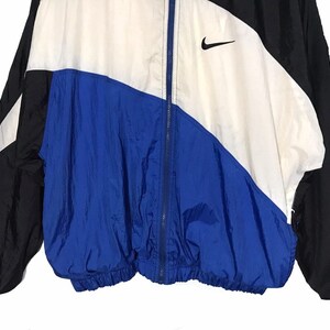 Op de afbeelding: Een blauw, wit en zwart windjack met een ritssluiting en een Nike-logo aan de rechterkant van de borst.