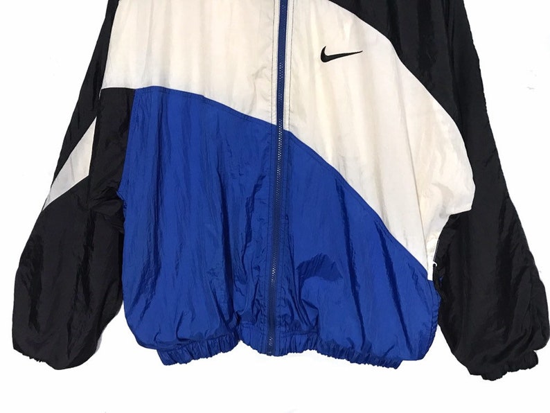 Op de afbeelding: Een blauw, wit en zwart windjack met een ritssluiting en een Nike-logo aan de rechterkant van de borst.