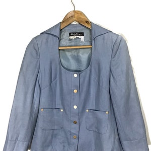 Puede incluir: Una chaqueta azul claro con cuello redondo, botones dorados y dos bolsillos de solapa. La chaqueta tiene una etiqueta que dice "Salvatore Ferragamo".