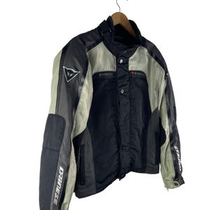 K&ouml;nnte beinhalten: Schwarze, graue und beige Motorradjacke mit dem Markennamen "DAINESE" auf den &Auml;rmeln. Die Jacke hat einen Stehkragen, mehrere Eins&auml;tze und ein dreieckiges Logo auf der Brust. Sie h&auml;ngt an einem Holzkleiderb&uuml;gel.