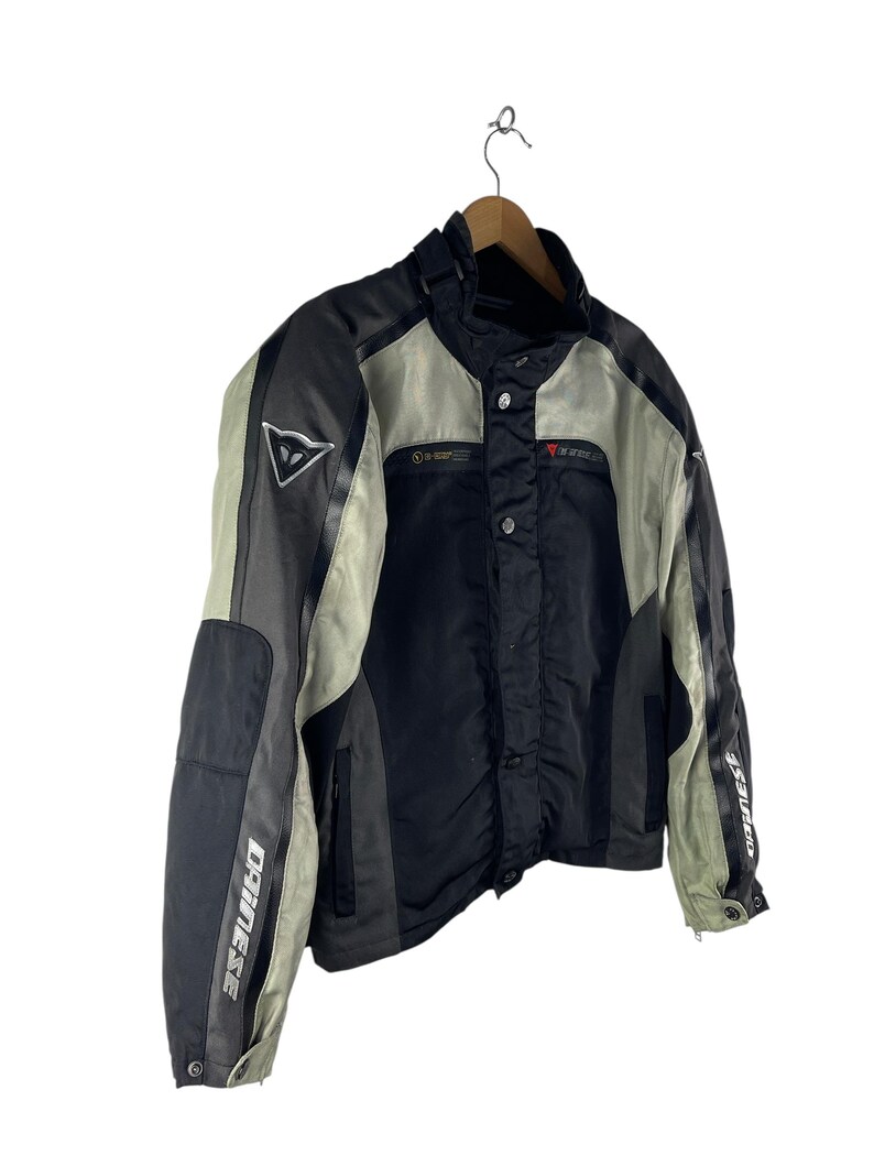 K&ouml;nnte beinhalten: Schwarze, graue und beige Motorradjacke mit dem Markennamen "DAINESE" auf den &Auml;rmeln. Die Jacke hat einen Stehkragen, mehrere Eins&auml;tze und ein dreieckiges Logo auf der Brust. Sie h&auml;ngt an einem Holzkleiderb&uuml;gel.