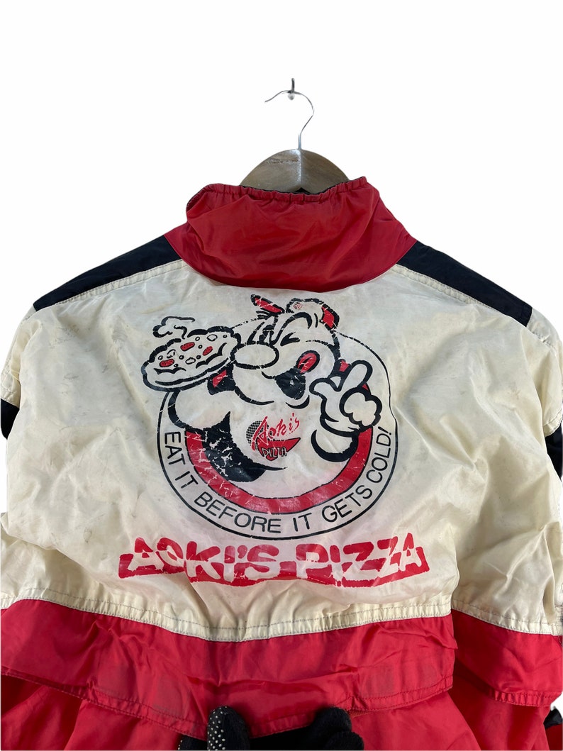 Puede incluir: Una chaqueta vintage blanca, roja y negra con un personaje de dibujos animados sosteniendo una pizza y el texto "Aoki's Pizza" y "Eat it before it gets cold!"
