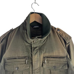 May include: Olive green military-style jacket with a ribbed collar and multiple pockets. The jacket has a zipper closure and decorative buttons. The jacket is hanging on a wooden hanger.