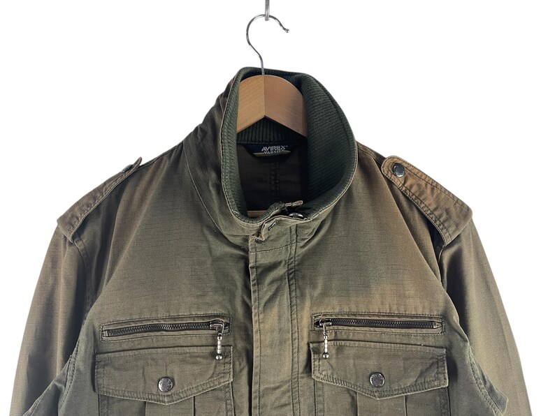 May include: Olive green military-style jacket with a ribbed collar and multiple pockets. The jacket has a zipper closure and decorative buttons. The jacket is hanging on a wooden hanger.