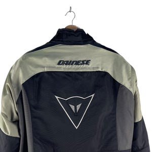 K&ouml;nnte beinhalten: Schwarze und graue Motorradjacke mit dem Markennamen "DAINESSE" auf den Schultern. Die Jacke hat ein wei&szlig;es Logo auf dem R&uuml;cken und h&auml;ngt an einem Kleiderb&uuml;gel.