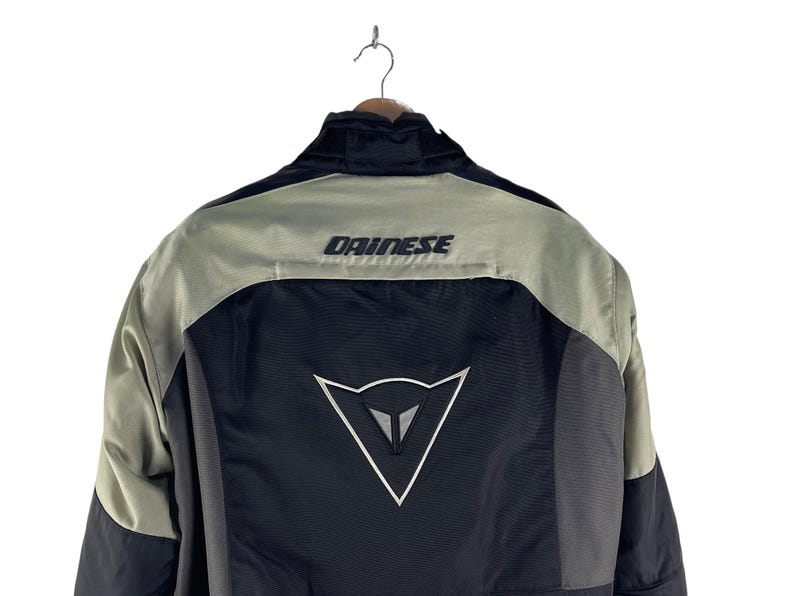 K&ouml;nnte beinhalten: Schwarze und graue Motorradjacke mit dem Markennamen "DAINESSE" auf den Schultern. Die Jacke hat ein wei&szlig;es Logo auf dem R&uuml;cken und h&auml;ngt an einem Kleiderb&uuml;gel.