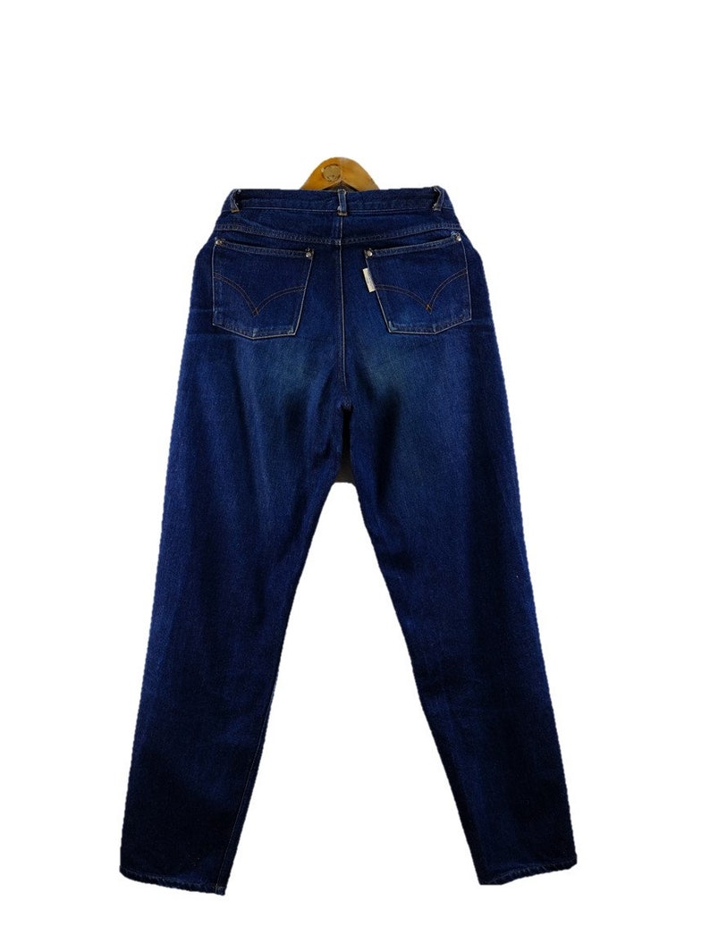 以下が含まれることがあります： ハイウエストでストレートレッグのダークブルーのデニムジーンズ。ジーンズは、クラシックな5ポケットのデザインとボタンフライです。