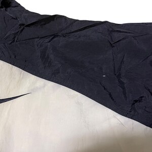 Op de afbeelding: Een zwart-witte windjack met een Nike swoosh-logo aan de linkerkant van de borst. De jas heeft een ritssluiting en een opstaande kraag.