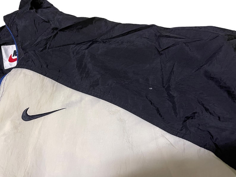 Op de afbeelding: Een zwart-witte windjack met een Nike swoosh-logo aan de linkerkant van de borst. De jas heeft een ritssluiting en een opstaande kraag.
