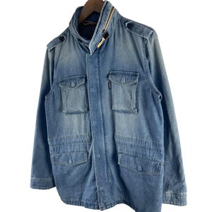 Puede incluir: Chaqueta vaquera azul claro con cuello alto y m&uacute;ltiples bolsillos delanteros. La chaqueta tiene cierre de cremallera y detalles de botones en los hombros. Cuelga de una percha de madera sobre fondo blanco.