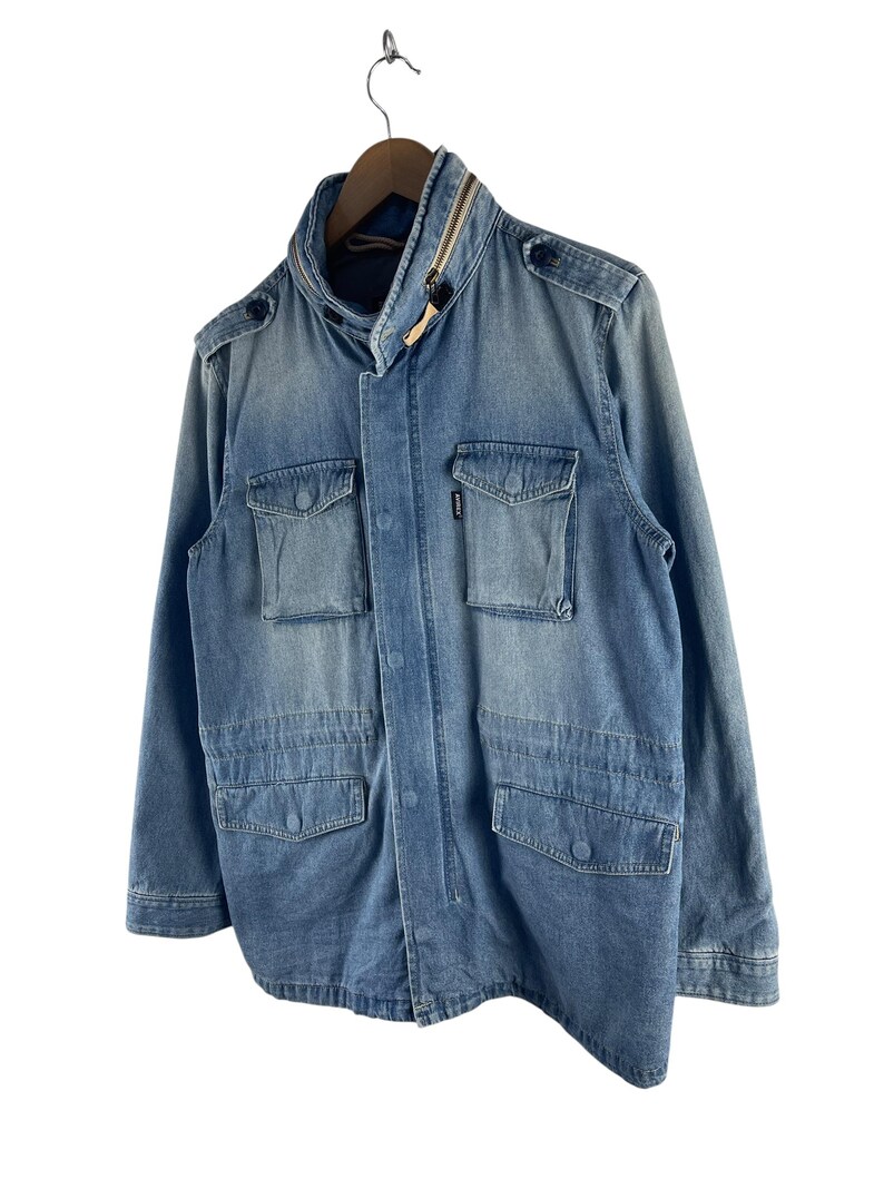 Puede incluir: Chaqueta vaquera azul claro con cuello alto y m&uacute;ltiples bolsillos delanteros. La chaqueta tiene cierre de cremallera y detalles de botones en los hombros. Cuelga de una percha de madera sobre fondo blanco.