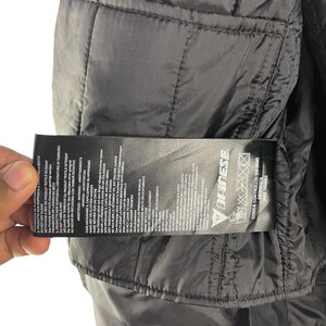 K&ouml;nnte beinhalten: Nahaufnahme des Innenraums einer schwarzen Steppjacke. Ein Etikett mit Pflegehinweisen und dem Markennamen "Dainese" ist sichtbar. Die Jacke hat einen Rei&szlig;verschluss und besteht aus einem glatten, gl&auml;nzenden Material.