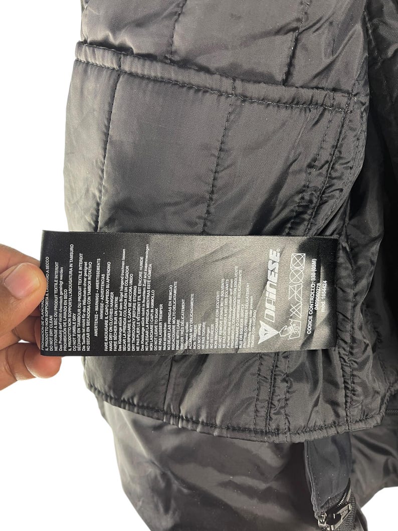 K&ouml;nnte beinhalten: Nahaufnahme des Innenraums einer schwarzen Steppjacke. Ein Etikett mit Pflegehinweisen und dem Markennamen "Dainese" ist sichtbar. Die Jacke hat einen Rei&szlig;verschluss und besteht aus einem glatten, gl&auml;nzenden Material.