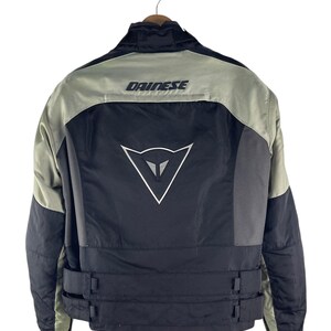 K&ouml;nnte beinhalten: Schwarze und graue Motorradjacke mit dem Markennamen "DAINESSE" auf den Schultern. Die Jacke hat ein wei&szlig;es Logo auf dem R&uuml;cken und verstellbare Riemen an der Taille. Die Jacke h&auml;ngt an einem Holzkleiderb&uuml;gel.