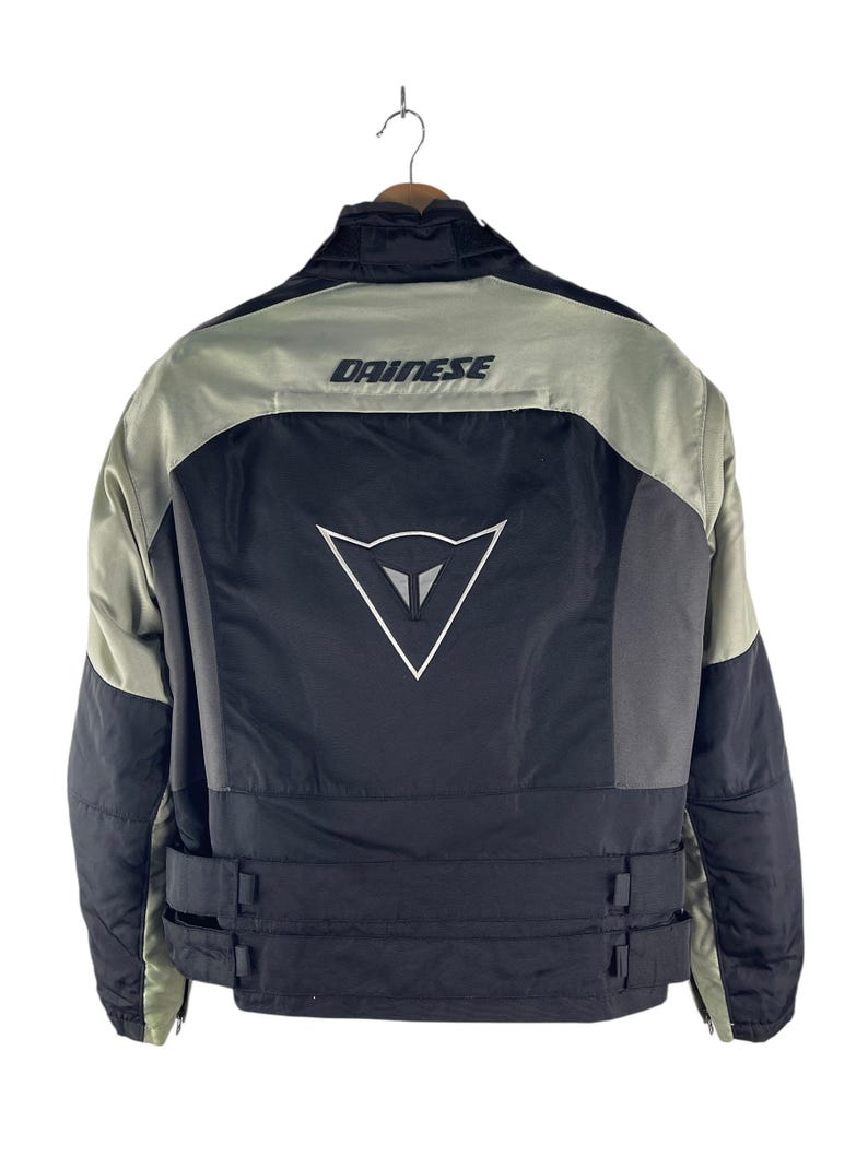K&ouml;nnte beinhalten: Schwarze und graue Motorradjacke mit dem Markennamen "DAINESSE" auf den Schultern. Die Jacke hat ein wei&szlig;es Logo auf dem R&uuml;cken und verstellbare Riemen an der Taille. Die Jacke h&auml;ngt an einem Holzkleiderb&uuml;gel.