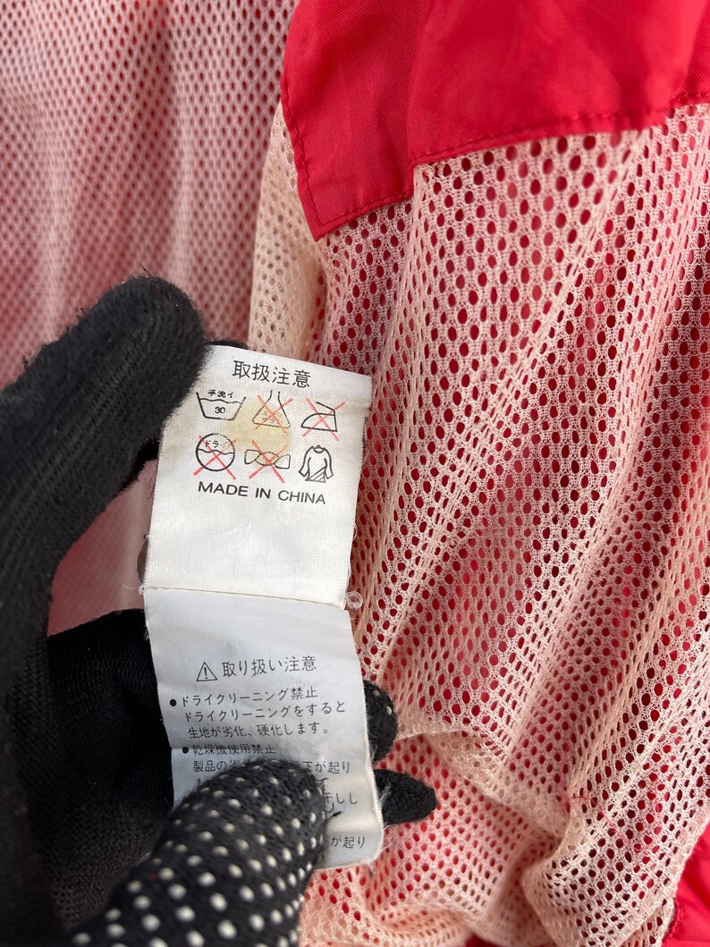 Puede incluir: Una etiqueta blanca con texto negro y s&iacute;mbolos, unida a una chaqueta roja y blanca. La etiqueta dice "取扱注意" (que significa "Precauciones de manejo") y "MADE IN CHINA". La etiqueta tambi&eacute;n incluye una serie de s&iacute;mbolos que indican las instrucciones de cuidado, como "30", "手洗い", "アイロン禁止" y "ドライクリーニング禁止".