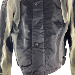 K&ouml;nnte beinhalten: Eine schwarze und graue Motorradjacke mit Knopfleiste. Die Jacke hat einen hohen Kragen und tr&auml;gt den Markennamen "Dainese" in Rot. Die Jacke hat schwarze Lederakzente und ein graues Futter.