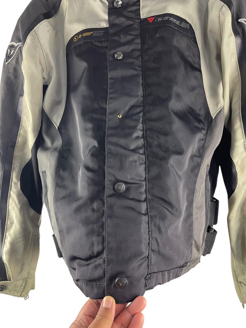 K&ouml;nnte beinhalten: Eine schwarze und graue Motorradjacke mit Knopfleiste. Die Jacke hat einen hohen Kragen und tr&auml;gt den Markennamen "Dainese" in Rot. Die Jacke hat schwarze Lederakzente und ein graues Futter.