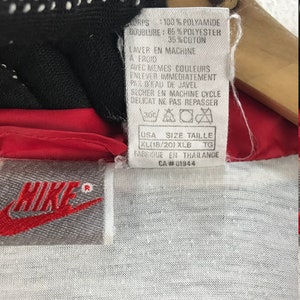 Puede incluir: Una chaqueta cortavientos Nike roja y blanca con una etiqueta blanca que dice "CORPS: 100% POLYAMIDE DOUBLURE: 65% POLYESTER 35%COTON LAVER EN MACHINE A FROID AVEC MEMES COULEURS ENLEVER IMMEDIATEMENT PAS D'EAU DE JAVEL SECHER EN MACHINE CYCLE DELICAT NE PAS REPASSER USA SIZE TAILLE XL(18/20) XLB TG FABRIQUE EN THAILANDE CA#01944"