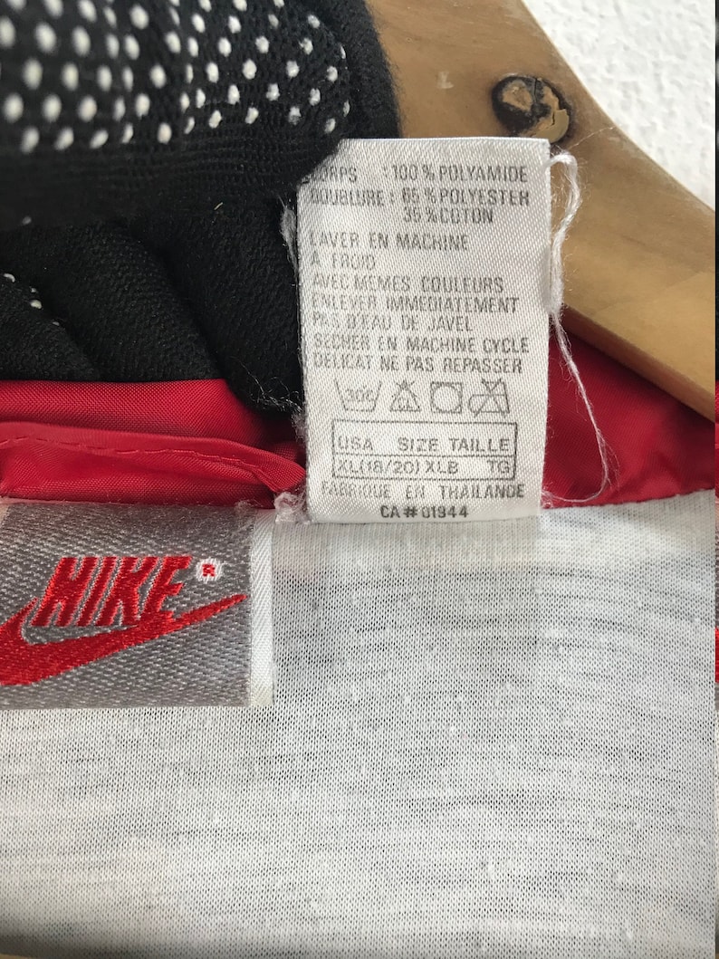 Puede incluir: Una chaqueta cortavientos Nike roja y blanca con una etiqueta blanca que dice "CORPS: 100% POLYAMIDE DOUBLURE: 65% POLYESTER 35%COTON LAVER EN MACHINE A FROID AVEC MEMES COULEURS ENLEVER IMMEDIATEMENT PAS D'EAU DE JAVEL SECHER EN MACHINE CYCLE DELICAT NE PAS REPASSER USA SIZE TAILLE XL(18/20) XLB TG FABRIQUE EN THAILANDE CA#01944"
